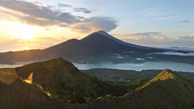 Mount Batur caldera and active volcano rise above Lake Batur with Mount Agung on the horizon at vibrant sunrise, lush green ridges, tranquil morning scene and dramatic light. Aerial view drone flight