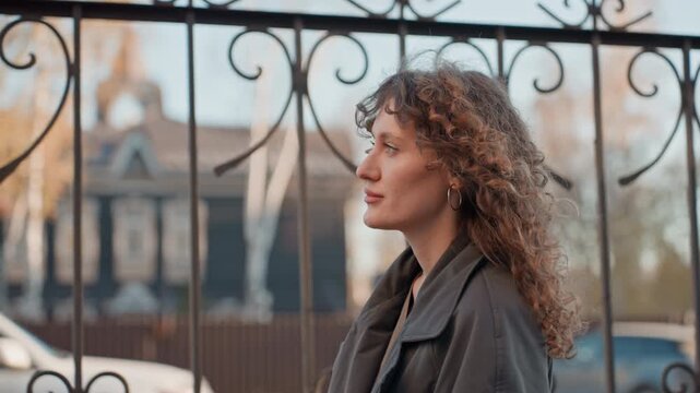 Closeup florist portrait by iron gate, curlyhaired woman smiles near ornate fence, soft daylight, urban storefront background, candid expression, poised before entering rustic shop, natural tones,