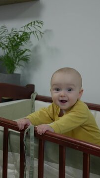 caucasian baby standing at crib smiling broadly, milestone achiever in yellow onesie showing joyful grin and playful gaze, bright nursery with potted plant and modern decor capturing