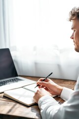 Fototapeta premium Side view man writing at desk near window with laptop