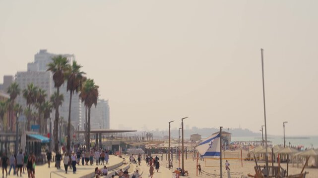 Tel Aviv promenade and beach as locals and tourists enjoy hot sunny day by sea. People swim walk and run by the Israeli coast on summer vacation holiday or weekend. Israeli flag waving in foreground