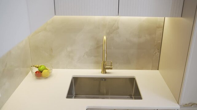 Smooth camera movement showing a modern kitchen interior. The shot focuses on a stylish white countertop, a stainless steel sink, and an elegant gold faucet with a marble backsplash