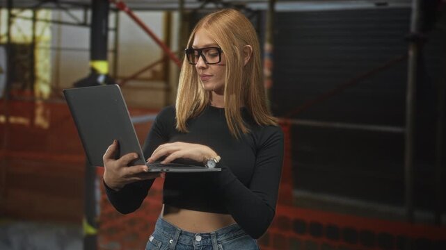 Woman holding laptop and typing, bare midriff visible, wearing glasses and jeans, standing amid scaffolding and safety netting in a building; concentration productivity.