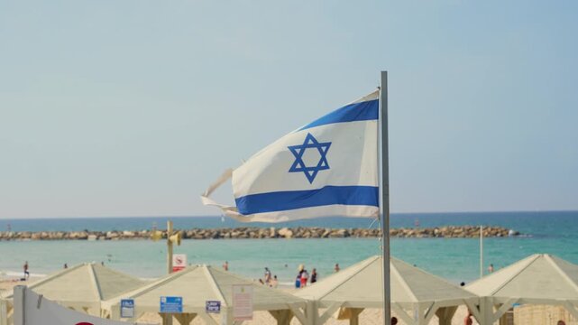 Close up on Israel flag waving in the wind by Tel Aviv beach during summer day. Hot weather by the Mediterranean Sea. Local people and tourists swimming and enjoying holiday vacation in Holy Land
