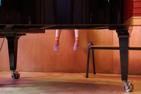 The feet of a school girl dangle behind a piano during a play recital.