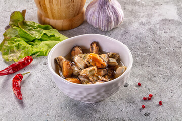 Fototapeta premium Marinated mussels in a white bowl with chili peppers, garlic, and fresh lettuce on a concrete background.