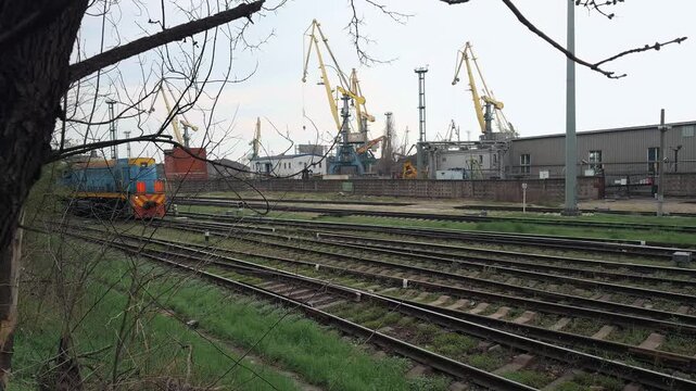 Authentic industrial diesel locomotive train moving on multiple railroad tracks through commercial sea port logistics zone during overcast daylight with harbor cranes in background wide angle shot