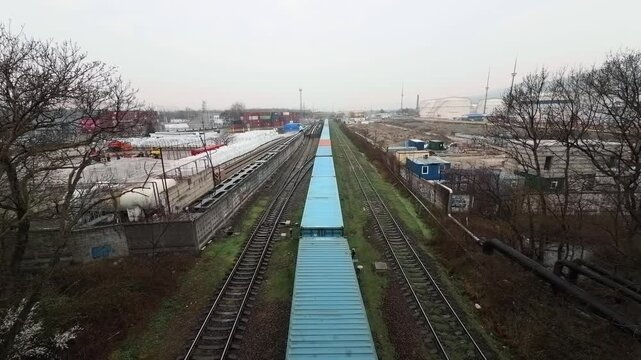 High angle ultra wide shot of long freight train with blue and red intermodal shipping containers moving on railroad tracks in industrial district of Novorossiysk during overcast day authentic