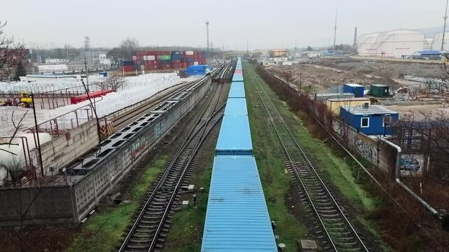 High angle perspective view of a long freight train transporting blue and red intermodal shipping containers on railroad tracks passing through industrial district in Novorossiysk authentic shot