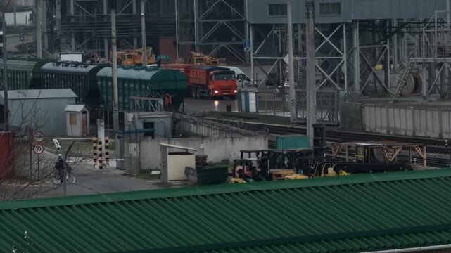 Professional port worker in safety vest standing on moving freight train wagon while cyclist waits at security barrier gate in Novorossiysk commercial sea port industrial zone authentic handheld shot