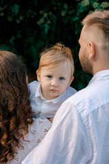 A young boy with blue eyes is held between his parents. The family stands outdoors against a green...