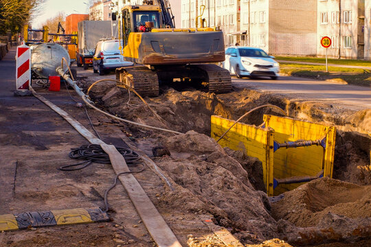 Excavator digging trench for underground utility repair on city street. Yellow trench shield box installed for worker safety. Road construction site with heavy machinery and pipes.