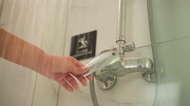 Hotel shower faucet being inspected, chrome fixture glistening with water droplets, flexible hose hanging against tiled wall, small hotel logo plaque visible, guest hand reaching to test temperature,