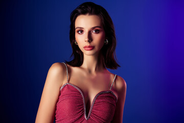 Fototapeta premium Stunning young woman in a stylish pink dress posing against a vibrant blue background with glamorous makeup and jewelry