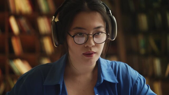 Korean female student in headphones having online video lesson, taking notes