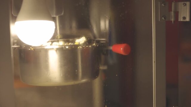 Close-up of a popcorn machine during popping. The machine is stainless steel, with a red handle