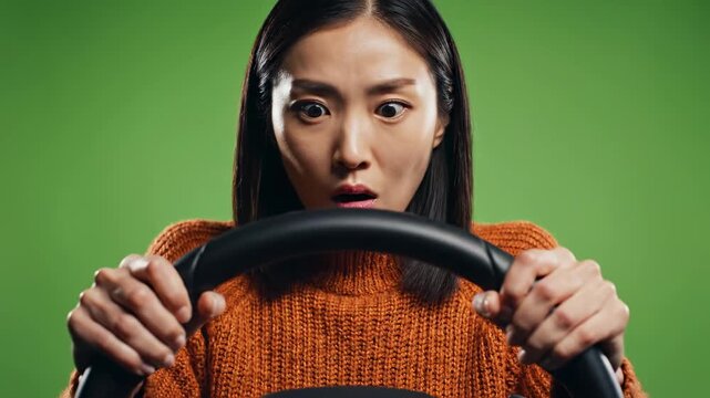 Asian Student Driver Gripping Steering Wheel, Learning Driving In Tense First Lesson Closeup Studio Shot With Green Background, Orange Sweater, Wide Eyes And Furrowed Brow, Tight Hands
