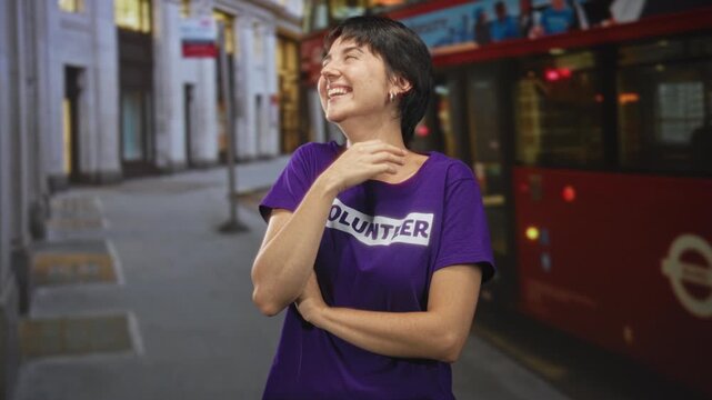 Young volunteer woman laughing with hand to mouth by red bus on street in a purple tshirt; community service joy.
