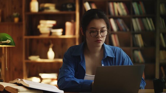 Korean female student does research in a library, types on laptop, reads a book