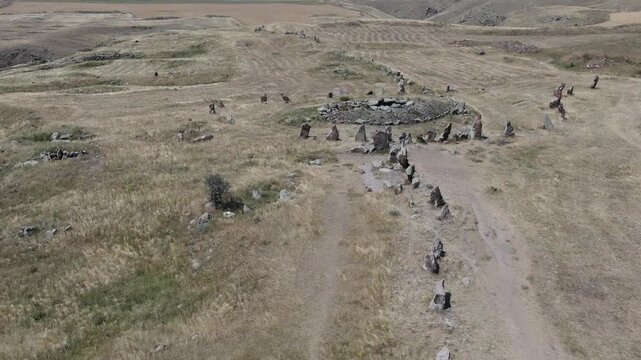 Aerial drone view flying over an ancient historical megalithic monument featuring a circle of standing stones and a central burial mound set in a dry, grassy rural landscape.