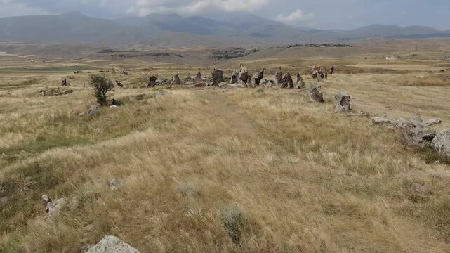 Aerial drone view flying over an ancient historical megalithic monument featuring a circle of standing stones and a central burial mound set in a dry, grassy rural landscape.