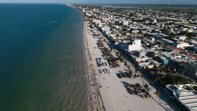 A dynamic aerial view of a lively beach where the natural rhythm of the sea meets organized human activity. Rows of umbrellas and palapas create geometric patterns on the sand, contrasted by the organ