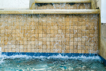 Clear water cascades down a tan square tiled wall into a blue rimmed basin creating splashing droplets under bright sunlight. © AncientAftertone