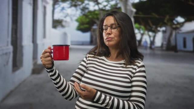 Woman in striped top holds red mug with left hand and gestures with right hand on a street near white buildings and trees; quiet contemplation.