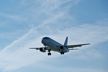 Obraz premium White passenger plane on final approach on a sunny day with blue sky, light clouds and beautiful backlighting. Sunlit white airplane landing against a blue sky with wispy clouds, backlit by bright sun