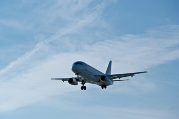 Obraz premium White passenger plane on final approach on a sunny day with blue sky, light clouds and beautiful backlighting. Sunlit white airplane landing against a blue sky with wispy clouds, backlit by bright sun