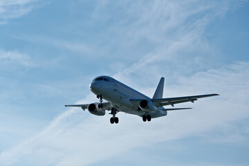 Fototapeta premium White passenger plane on final approach on a sunny day with blue sky, light clouds and beautiful backlighting. Sunlit white airplane landing against a blue sky with wispy clouds, backlit by bright sun