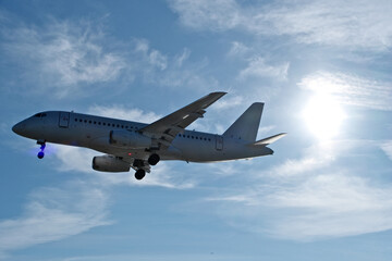 Obraz premium White passenger plane on final approach on a sunny day with blue sky, light clouds and beautiful backlighting. Sunlit white airplane landing against a blue sky with wispy clouds, backlit by bright sun