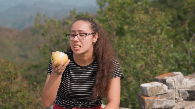 Multiracial woman eating apple on stone wall, closeup portrait with curly hair and glasses, dotted shirt and casual jeans, quiet hillside view, student daydreaming then checking map, solo hiker taking