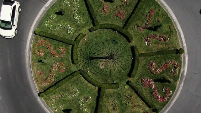 Overhead aerial view of a busy city traffic roundabout. Cars and a bus navigate the circular intersection featuring a green center, surrounded by urban buildings, trees, and sidewalks.