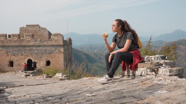 Young woman backpacker resting on ruins with apple, stone fortress in background, wide mountain vista, quiet reflective mood, casual striped shirt and sneakers, red jacket tied at waist, light breeze