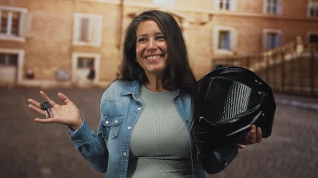 Woman holding motorcycle helmet and keys smiles and gestures with open hand in street by a building; joy adventure.