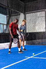 Obraz premium Diverse male partners playing paddle sport on blue court indoors holding paddles and yellow balls
