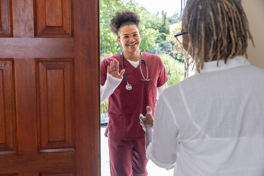 Mid adult nurse in maroon scrubs with stethoscope greeting senior African American woman at doorway