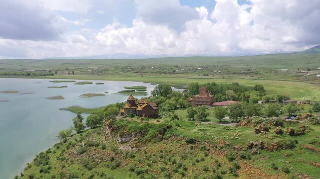 A drone flies backward over a rocky, grassy hill to reveal a beautiful ancient stone monastery complex overlooking a vast, calm blue lake and distant majestic mountain landscape.
