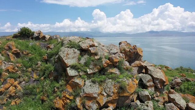 A drone flies backward over a rocky, grassy hill to reveal a beautiful ancient stone monastery complex overlooking a vast, calm blue lake and distant majestic mountain landscape.