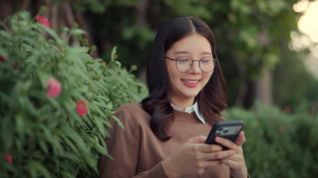 Happy young Asian woman wearing glasses smiling while using a smartphone in a lush green garden during the day, representing digital connectivity, social media engagement, and a balanced lifestyle