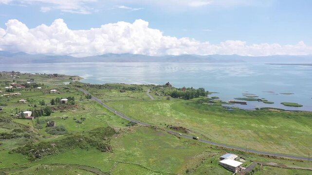 A drone flies backward over a rocky, grassy hill to reveal a beautiful ancient stone monastery complex overlooking a vast, calm blue lake and distant majestic mountain landscape.