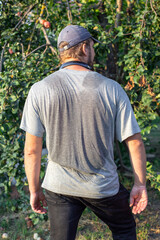Fototapeta premium Man's back in a grey t-shirt and jeans, wearing a cap and standing outdoors. Gardening, summer day, casual attire.