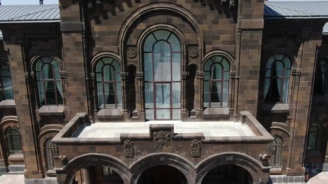 Drone footage flying towards the grand Gevorgian Seminary building within the Mother See of Holy Etchmiadzin complex in Armenia, showcasing its beautiful dark stone architecture and surrounding garden