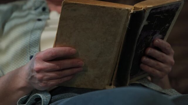 Woman reading old hardback book while sitting on sofa in dimly lit room close-up