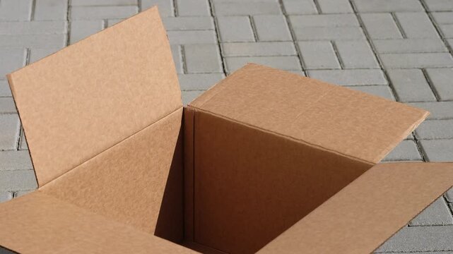 An empty open cardboard box sitting on a grey paved outdoor surface with its flaps fluttering in the wind. Close up.