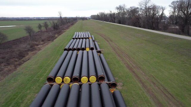 Rows of long cylindrical steel pipes with dark protective coating and yellow end caps stacked neatly on a grassy field