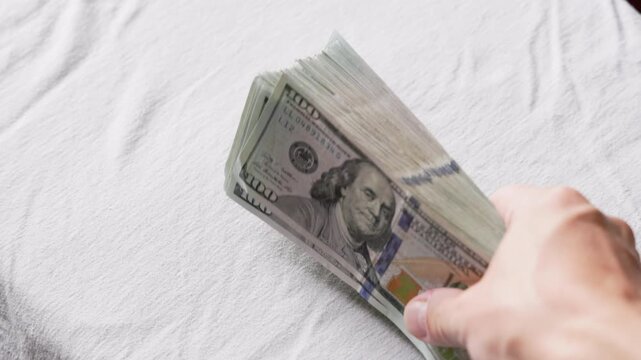 Person's hand displays a large stack of hundred-dollar bills on textured white surface. Scene evokes feelings of wealth and financial success.