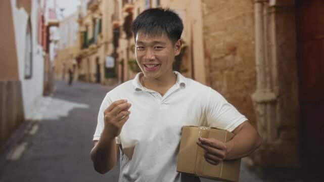 Man smiling, holding cardboard box and gesturing with fingers on a narrow stone street; helpful delivery service.