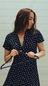 Caucasian woman inspecting hanger before choosing outfit in quiet fitting room, thoughtful pause, close study of hook and fabric, mirror reflection, retail rack at edge, deliberate wardrobe decision